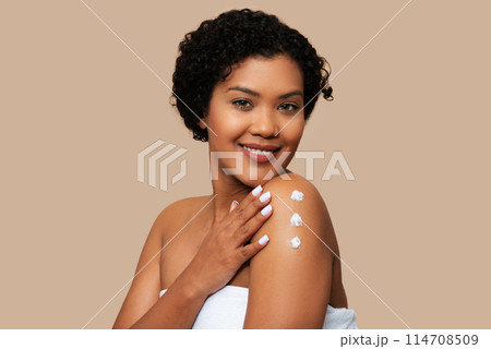 A cheerful young woman is captured as she gently applies cream to her shoulder, showcasing her skincare routine. Her radiant smile and clear complexion suggest a sense of well-being A cheerful young woman is captured as she gently applies cream to her shoulder, showcasing her skincare routine. Her radiant smile and clear complexion suggest a sense of well-being 114708509