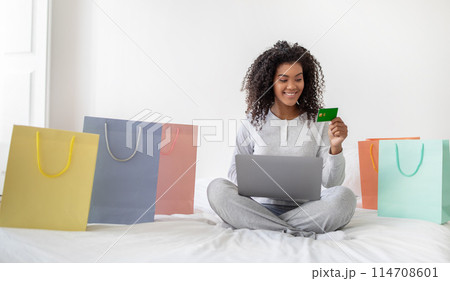 A cheerful young Hispanic woman is seated on bed with her legs crossed, surrounded by colorful shopping bags, holding a credit card in hand while looking at her laptop screen A cheerful young Hispanic woman is seated on bed with her legs crossed, surrounded by colorful shopping bags, holding a credit card in hand while looking at her laptop screen 114708601