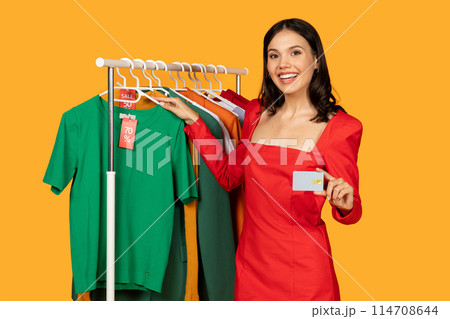 A woman is standing in front of a rack of clothes, holding a credit card in her hand. She appears to be shopping for clothing items at a store, making a payment with the credit card. A woman is standing in front of a rack of clothes, holding a credit card in her hand. She appears to be shopping for clothing items at a store, making a payment with the credit card. 114708644