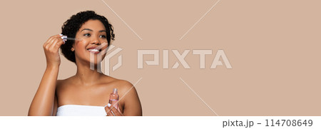 A smiling young woman with her hair up is applying a clear facial serum to her cheek using a dropper. She is wrapped in a white towel, indicating morning routine, panorama, copy space A smiling young woman with her hair up is applying a clear facial serum to her cheek using a dropper. She is wrapped in a white towel, indicating morning routine, panorama, copy space 114708649