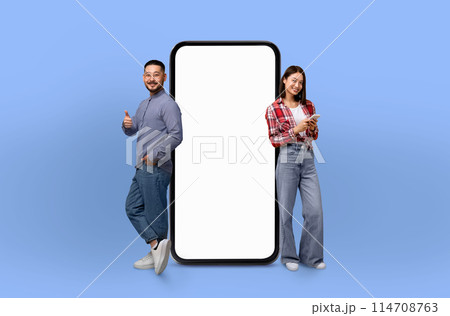 Asian couple standing next to a large-sized telephone, looking at the device with curiosity. The phone appears oversized in comparison to the people, creating a unique visual contrast. Asian couple standing next to a large-sized telephone, looking at the device with curiosity. The phone appears oversized in comparison to the people, creating a unique visual contrast. 114708763