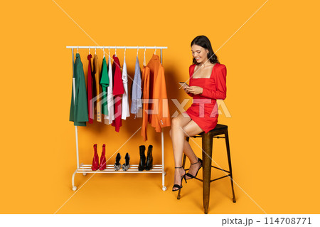 A stylish woman sits on a black stool, wearing a vibrant red dress that contrasts with the yellow background. She is engrossed in her smartphone, possibly scrolling through a shopping app 114708771