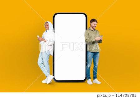 European man and black woman in hijab standing side by side, both looking at a cell phone in their hands, smartphone with white blank screen between them 114708783
