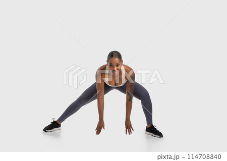 African American woman is seen performing a squat exercise on a white background. She is bending her knees while keeping her back straight, engaging her leg muscles. The white background 114708840