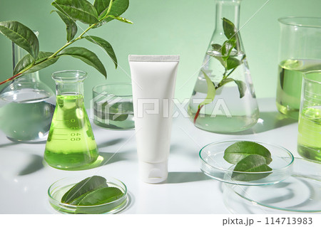 The sample tube of cosmetic without label displayed in center, petri dish with green tea leaves, an erlenmeyer of green tea extract and other experiment utensils in back. Mock up for advertising The sample tube of cosmetic without label displayed in center, petri dish with green tea leaves, an erlenmeyer of green tea extract and other experiment utensils in back. Mock up for advertising 114713983