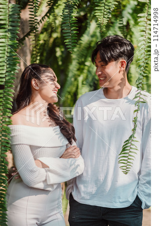 A couple standing in a lush green forest, smiling at each other. The woman is wearing a white shirt and the man is wearing a white shirt and jeans 114714998