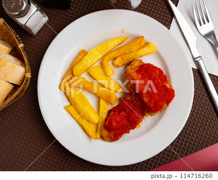 There is large slice of fried cod on plate, covered with tomato sauce and French fries. 114716260