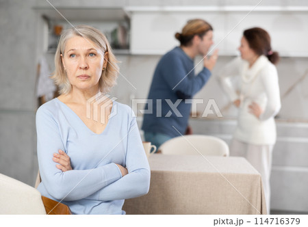 Upset old woman sitting in kitchen and man and woman arguing in background 114716379
