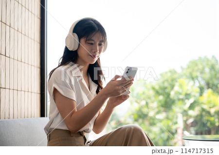 A beautiful Asian woman is using her smartphone while listening to music on her headphones in a cafe A beautiful Asian woman is using her smartphone while listening to music on her headphones in a cafe 114717315
