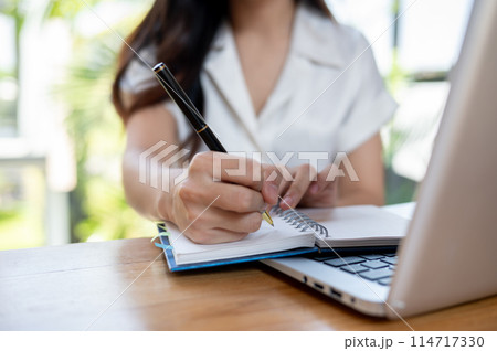 An Asian woman is listing her ideas in a notebook while browsing the internet on her laptop. An Asian woman is listing her ideas in a notebook while browsing the internet on her laptop. 114717330
