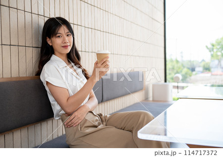 An attractive Asian woman sits in a coffee shop, a coffee cup in her hand, and smiles at the camera. An attractive Asian woman sits in a coffee shop, a coffee cup in her hand, and smiles at the camera. 114717372