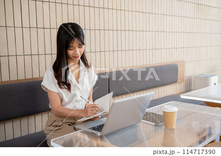 A businesswoman is working remotely from a coffee shop, working on her laptop and taking notes. 114717390