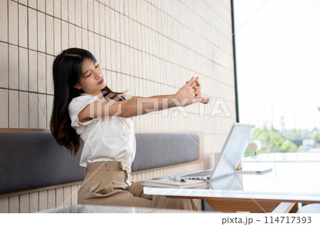 A beautiful Asian woman is feeling tired, stretching her hands while working remotely at a cafe. A beautiful Asian woman is feeling tired, stretching her hands while working remotely at a cafe. 114717393
