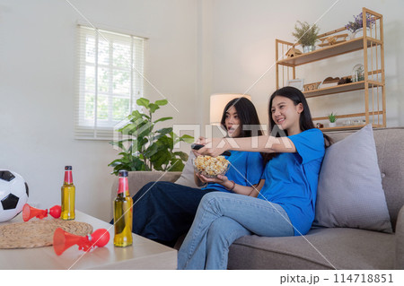 Lesbian couple cheering for Euro football at home with snacks and beer. Concept of LGBTQ pride, sports enthusiasm, and domestic leisure Lesbian couple cheering for Euro football at home with snacks and beer. Concept of LGBTQ pride, sports enthusiasm, and domestic leisure 114718851