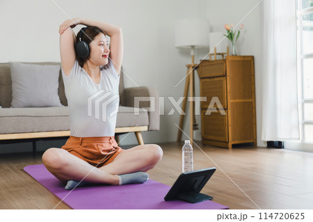Young Asian woman at home doing exercise in front of open laptop, repeating instructions by professional online fitness trainer. 114720625