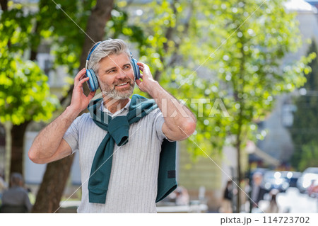 Happy mature man in wireless headphones choosing, listening music in smartphone application outdoors 114723702