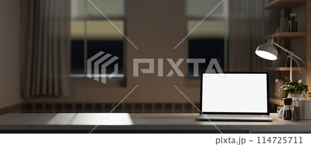 A home office workspace with a white-screen laptop computer mockup on a table in the evening. 114725711