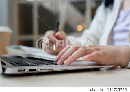 A woman working remotely at a cafe, using her laptop computer and taking notes in a book. A woman working remotely at a cafe, using her laptop computer and taking notes in a book. 114725756