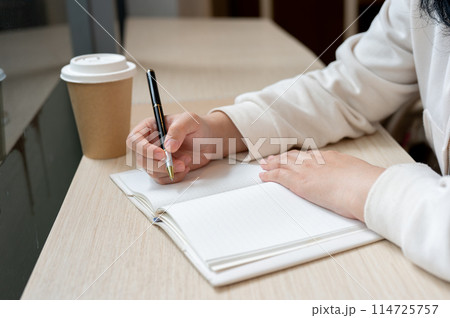 A cropped image of a woman sits in a coffee shop, writing something in a book or keeping her diary. A cropped image of a woman sits in a coffee shop, writing something in a book or keeping her diary. 114725757