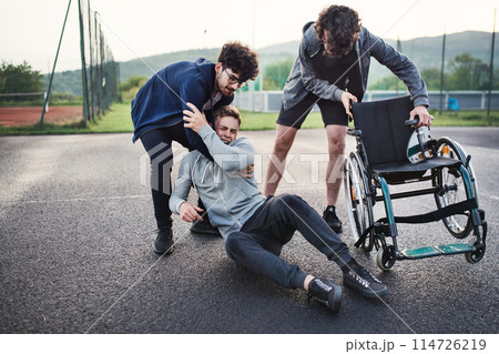 Young disabled man fall off wheelchair, fiends helping to sit back. Male friendship. 114726219