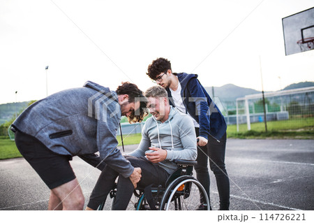 Young man in wheelchair spending free time outdoor with friends. Friends helping disabled man to sit in wheelchair. Male friendship. Young man in wheelchair spending free time outdoor with friends. Friends helping disabled man to sit in wheelchair. Male friendship. 114726221