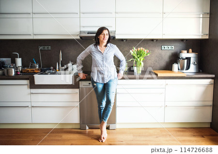Portrait of beautiful mature woman with black hair and soft smile, standing barefoot in kitchen. 114726688