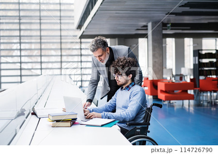 Handsome university professor helping student in wheelchair, showing him book for his thesis in library. 114726790