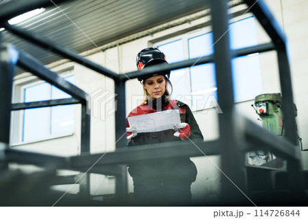 Beautiful blonde woman works as a welder in workshop, reading blueprints, wearing protective clothing and a welding mask. 114726844