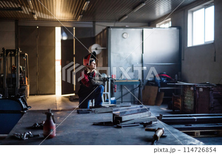 Beautiful blonde woman works as a welder in workshop, operating welding machine, wearing protective clothing and a welding mask. Beautiful blonde woman works as a welder in workshop, operating welding machine, wearing protective clothing and a welding mask. 114726854