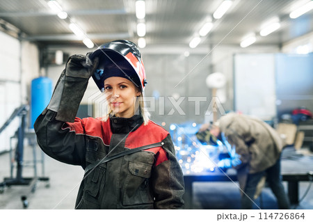 Beautiful blonde woman works as a welder in workshop, operating welding machine, wearing protective clothing and a welding mask. Beautiful blonde woman works as a welder in workshop, operating welding machine, wearing protective clothing and a welding mask. 114726864