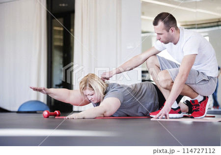 Overweight woman exercising on gym mat in gym. Personal trainer couching her and helping her. Overweight woman exercising on gym mat in gym. Personal trainer couching her and helping her. 114727112