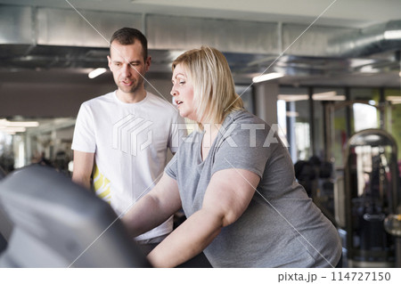 Overweight woman exercising on treadmill in gym. Personal trainer helping her. Overweight woman exercising on treadmill in gym. Personal trainer helping her. 114727150