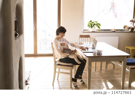 Mother with small baby working from homeoffice, typing on laptop. Mom paying bills online, holding small baby. 114727661