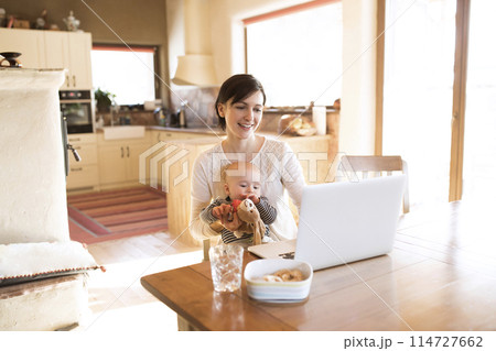 Mother with small baby working from homeoffice, typing on laptop. Mom paying bills online, holding small baby. 114727662
