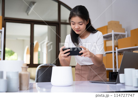Young Asian business entrepreneur using a smartphone take a photo of product for upload to website online shop. Young Asian business entrepreneur using a smartphone take a photo of product for upload to website online shop. 114727747