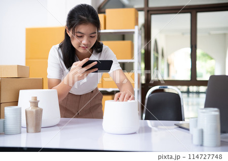 Young Asian business entrepreneur using a smartphone take a photo of product for upload to website online shop. Young Asian business entrepreneur using a smartphone take a photo of product for upload to website online shop. 114727748