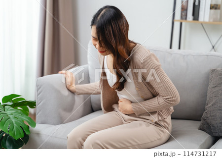The woman with a stomachache is holding her stomach in pain, sitting on the sofa in the living room. 114731217