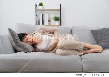 The woman with a stomachache is holding her stomach in pain, laying on the sofa in the living room. 114731219