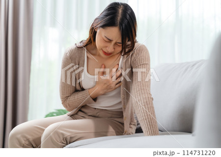 A woman is sitting on a couch with her hand holding on her chest, suffering from a heart attack. 114731220