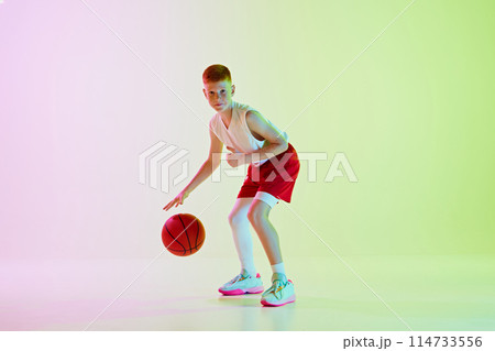 Focused boy, basketball player training, playing, dribbling ball against gradient background in neon light 114733556