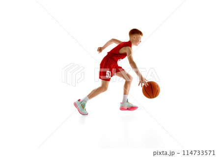 Dribbling skills. Active 11 years old boy, basketball player in red uniform in motion with ball, training isolated on white studio background Dribbling skills. Active 11 years old boy, basketball player in red uniform in motion with ball, training isolated on white studio background 114733571