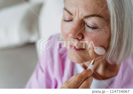 Skincare for mature woman. Older woman using facial roller for blood circulation, after serum. 114734067