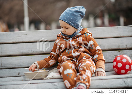 Cute toddler girl eating lunch in park, sitting on bench. Picnic in park during cold spring day. 114734314