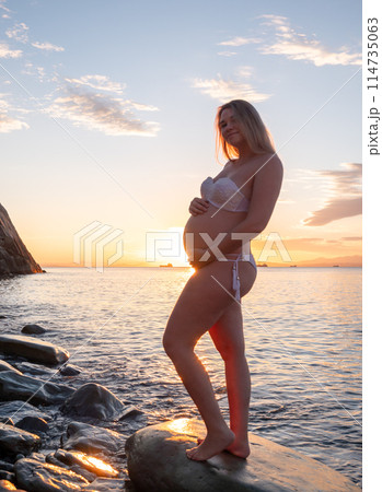 Pregnant woman in bikini posing on rocky beach at sunrise with mountain view 114735063