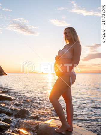 Pregnant woman in bikini posing on rocky beach at sunrise with mountain view 114735070