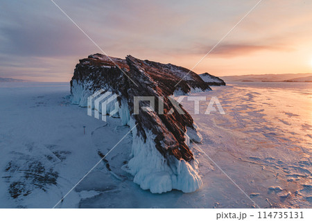 Aerial over the rocky island in lake Baikal. Winter landscape of frozen Baikal at beautiful orange sunrise. Sun reflections on the ice. Popular tourist spot 114735131