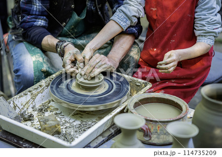 Potter teaching child to work on potter wheel, how to sculp clay pot. Closeup of dirty hands 114735209