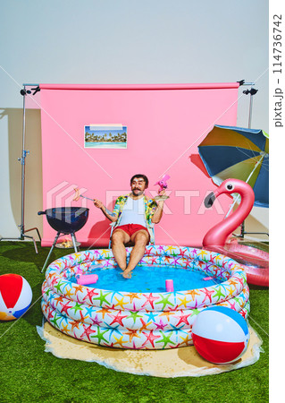 Young man in vibrant outfit with tropical shirt relaxing in inflatable swimming pool and cooking barbeque-sausages. 114736742