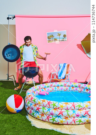 Overjoyed man in colorful, tropical shirt cooking barbeque, sausages in studio with pink background and kiddies swimming pool. 114736744