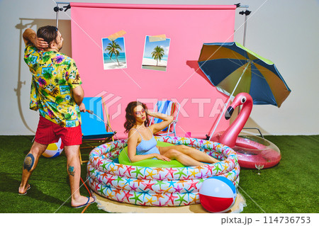 Young man spraying water from hose at beautiful woman in vibrant kiddie pool. Fun and festive summer vibe. 114736753
